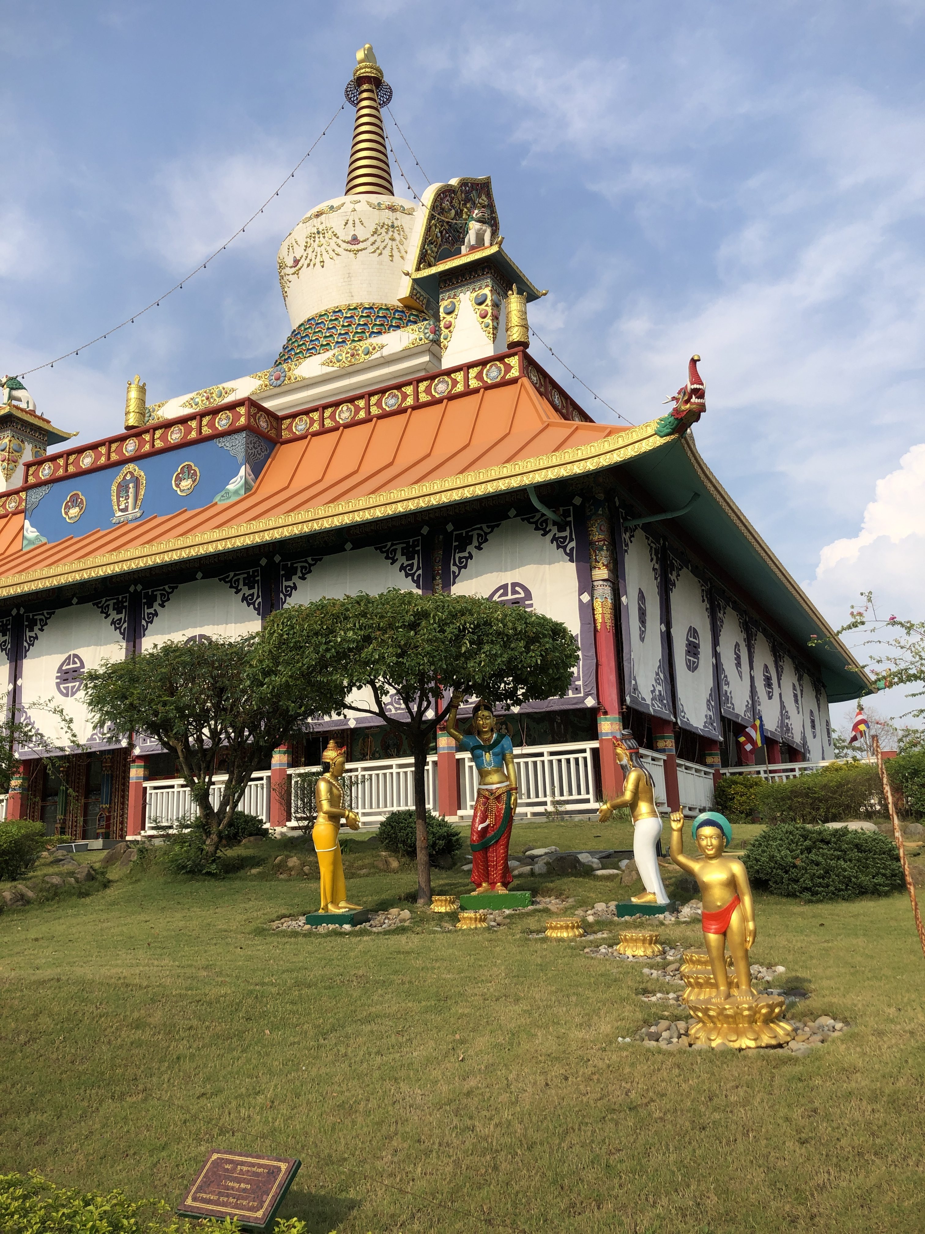 A colorful Buddhist temple with ornate architectural details, featuring a large stupa and statues of figures in traditional attire on a neatly maintained grassy area. The sky is clear and sunny.
