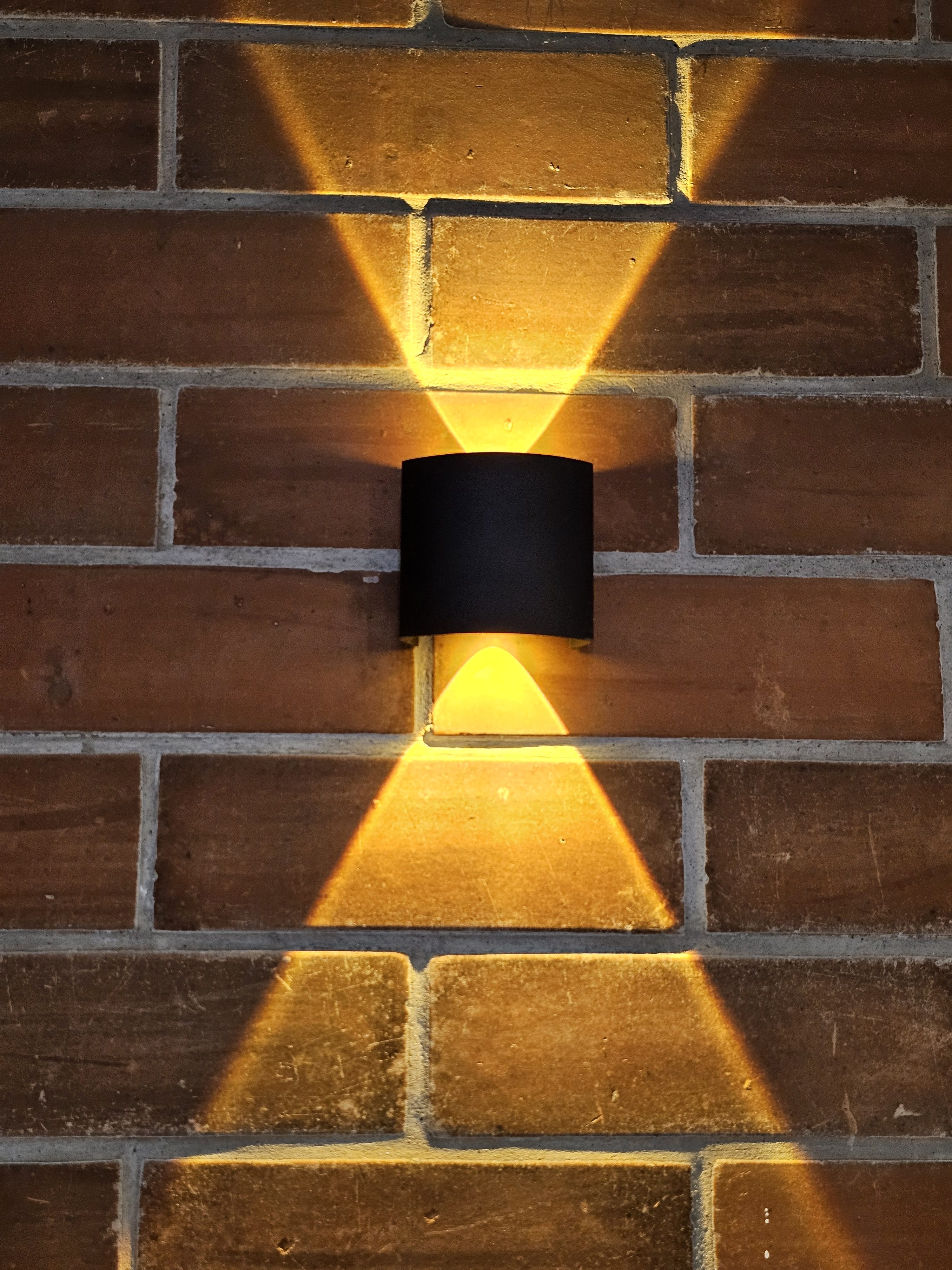 A black, cylindrical LED wall lamp mounted on a brick wall, emitting beams of warm light upwards and downwards, forming two inverted triangular shapes. The contrast between the light and shadow emphasizes the texture of the brickwork, creating a visually appealing pattern. Spotted it at a restaurant in Kozhikode, Kerala.