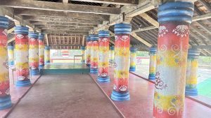 The image shows the interior of a wooden structure supported by several colorful columns. Each column features a unique design with vibrant red, yellow, and blue paint, depicting floral patterns and symbols. The wooden ceiling has exposed beams, and the floor is made of red tiles. There is a raised platform in the center of the room, surrounded by the ornate columns, creating an inviting and bright atmosphere. Natural light shines through openings, illuminating the space.