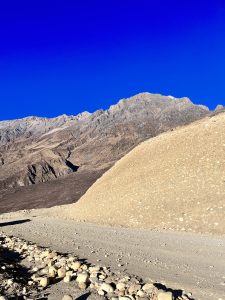 

A  mustang mountainous landscape under a clear blue sky, featuring rocky terrain and a dirt path in the foreground. A large slope with small rocks is prominent on the right, and rugged peaks are visible in the background