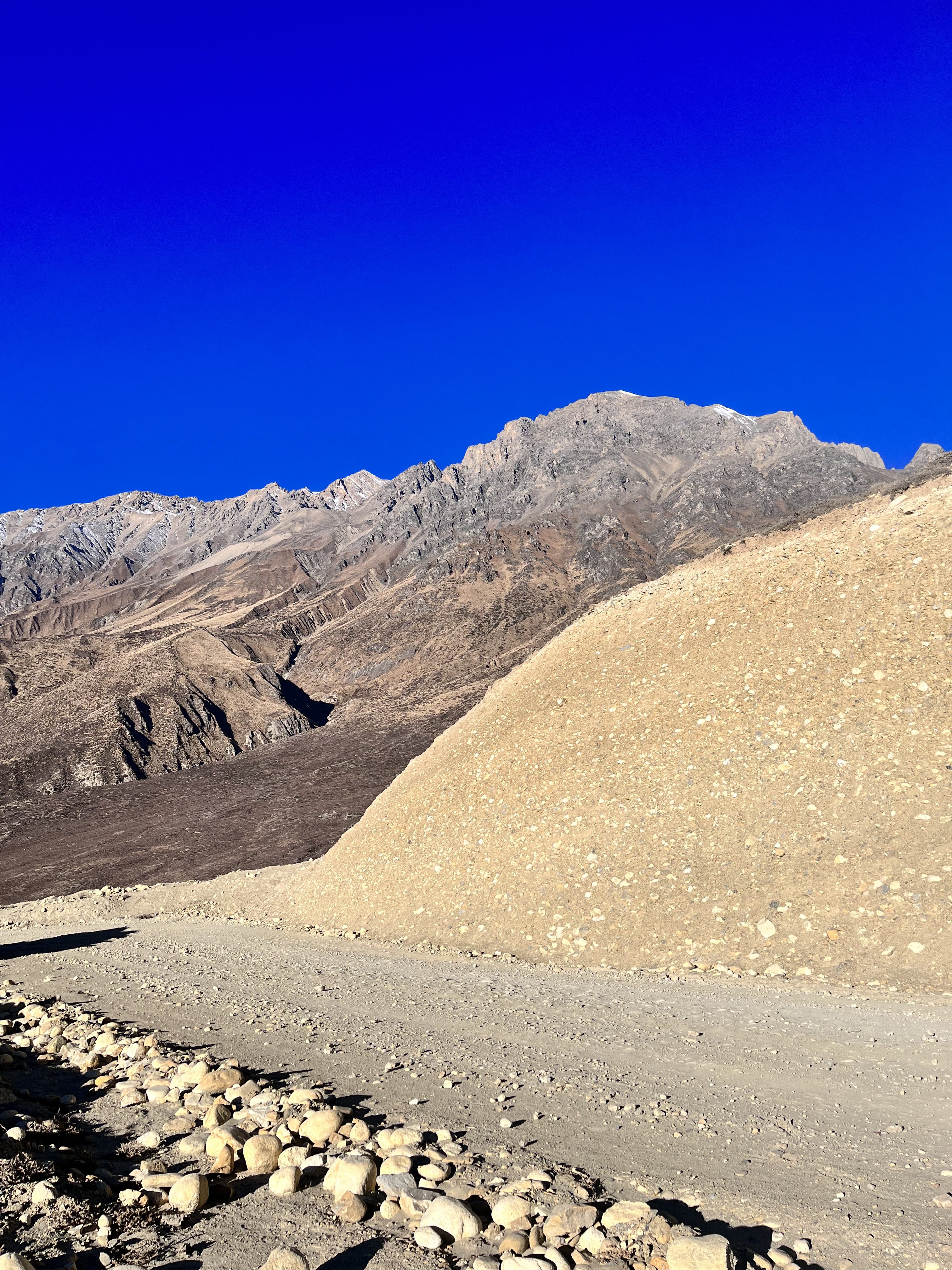 A mustang mountainous landscape under a clear blue sky, featuring rocky terrain and a dirt path in the foreground. A large slope with small rocks is prominent on the right, and rugged peaks are visible in the background