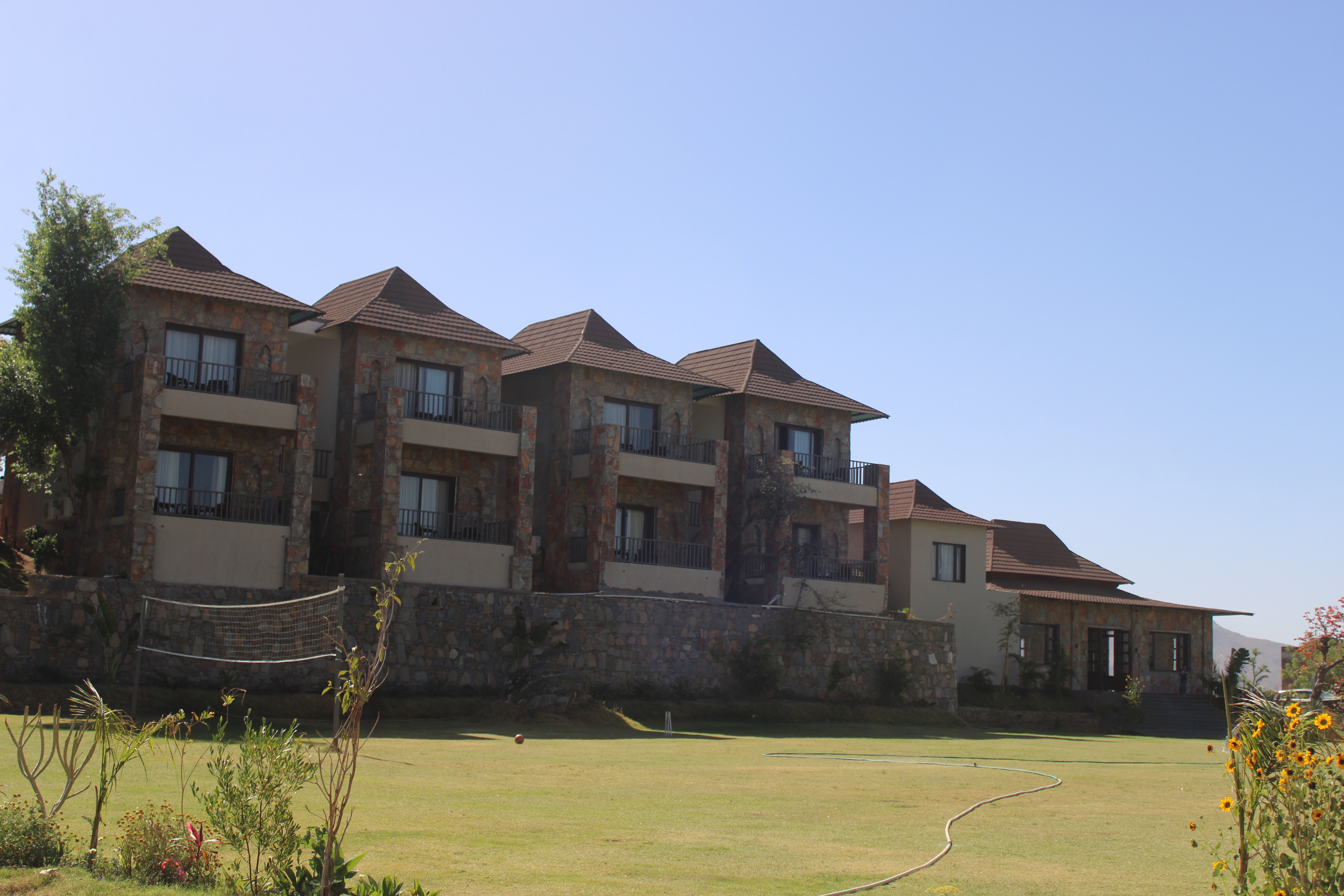 A residential building with stone exterior and multiple balconies, surrounded by a manicured lawn with a volleyball net and flowering plants under a clear blue sky.