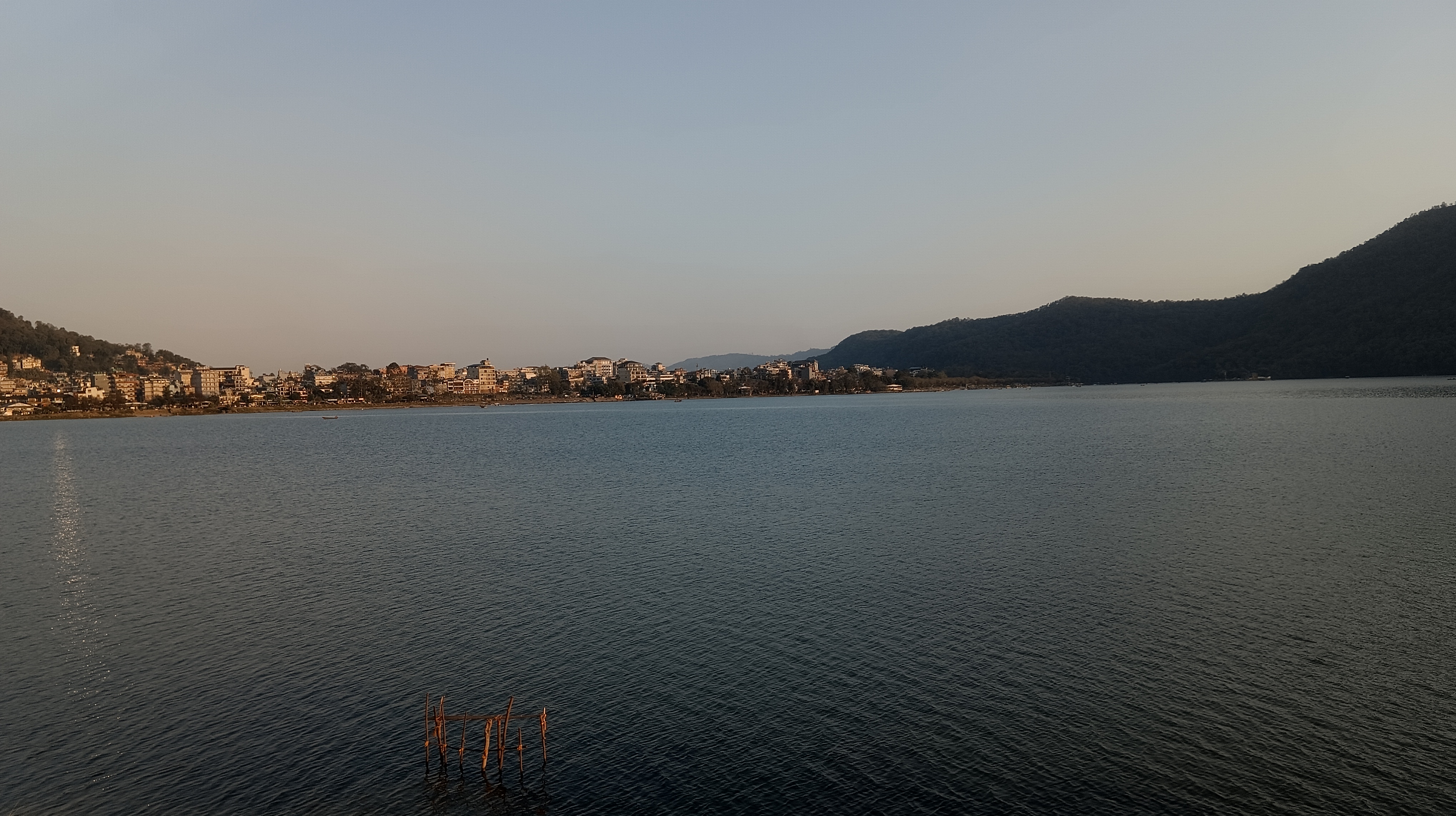 A panoramic view of a Fewa lake with a distant town along the shoreline, surrounded by hills under a clear sky.