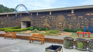 A large wall mural depicting historical figures and scenes, featuring soldiers and a person on horseback. The artwork includes elements like waving flags and faces, with the words "Philippine Veterans Legion" visible. In front of the mural are landscaped gardens with plants, wooden benches, and a small playground area with colorful play structures under a cloudy sky.