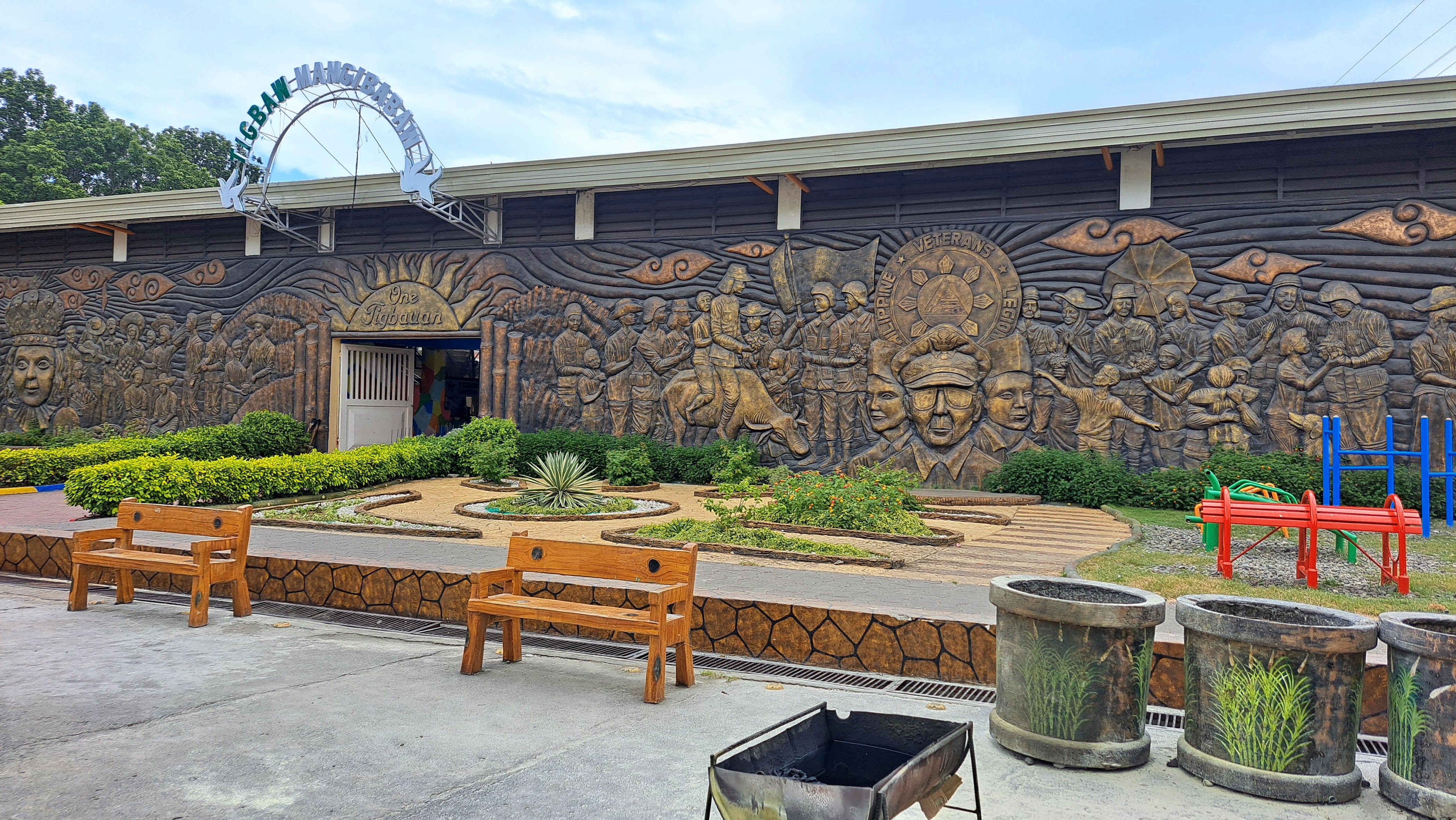 A large wall mural depicting historical figures and scenes, featuring soldiers and a person on horseback. The artwork includes elements like waving flags and faces, with the words "Philippine Veterans Legion" visible. In front of the mural are landscaped gardens with plants, wooden benches, and a small playground area with colorful play structures under a cloudy sky.