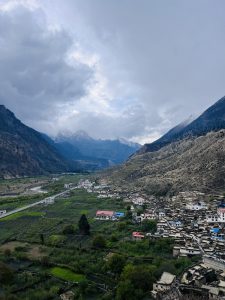 A scenic mountain valley with a cluster of houses nestled along the hillside, surrounded by green fields and distant snow-capped peaks under a cloudy sky.