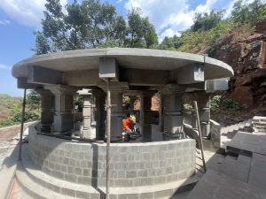 A partially built circular stone pavilion with ornate columns and a flat roof, nestled in a hilly landscape. At the center of the structure stands a stone sculpture of a bull. The backdrop features lush green trees, rugged terrain, and a clear blue sky.