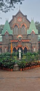 A Gothic-style stone building with pointed arches and green roofs, featuring symmetrical windows. In front, there’s a garden with potted plants and a white bust statue on a pedestal, accompanied by a yellow flag.
