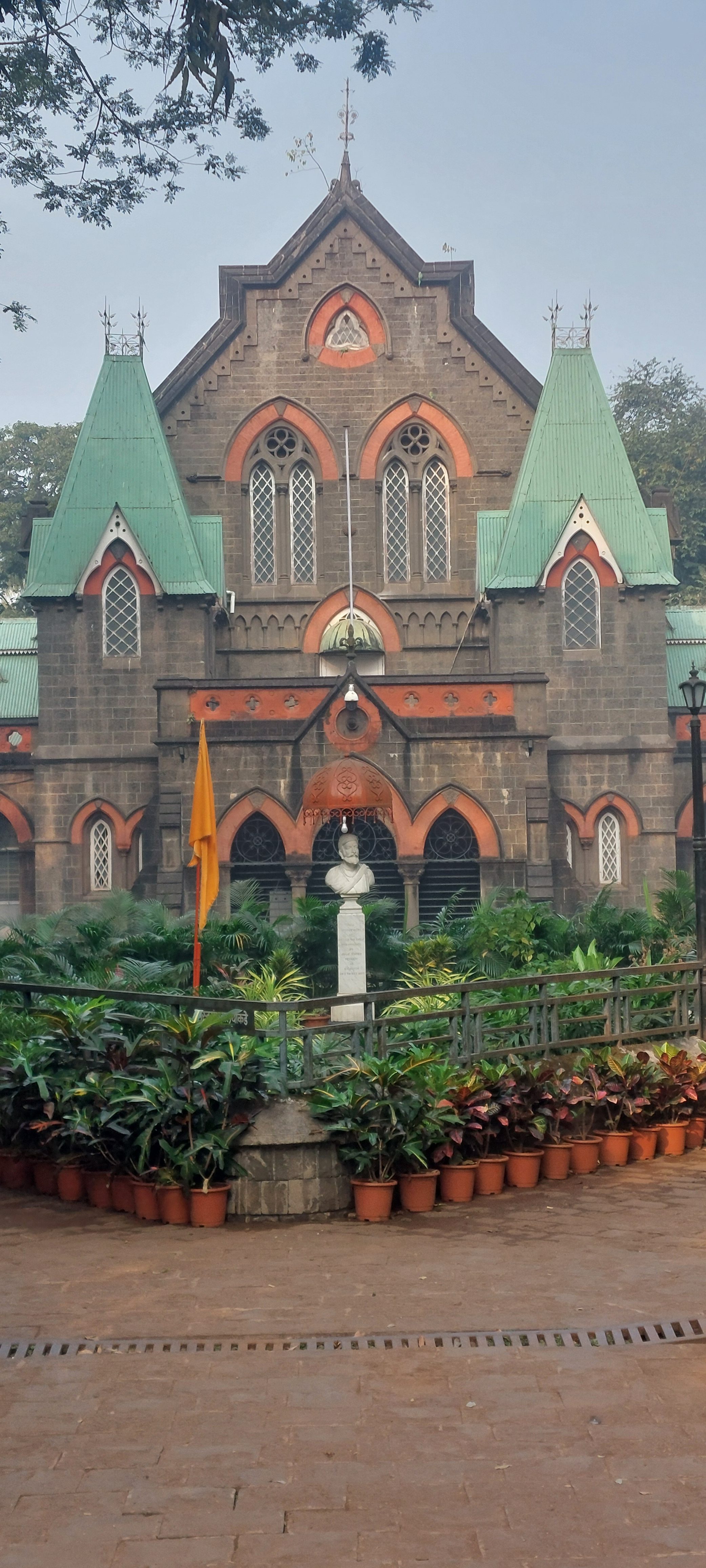 A Gothic-style stone building with pointed arches and green roofs, featuring symmetrical windows. In front, there's a garden with potted plants and a white bust statue on a pedestal, accompanied by a yellow flag.
