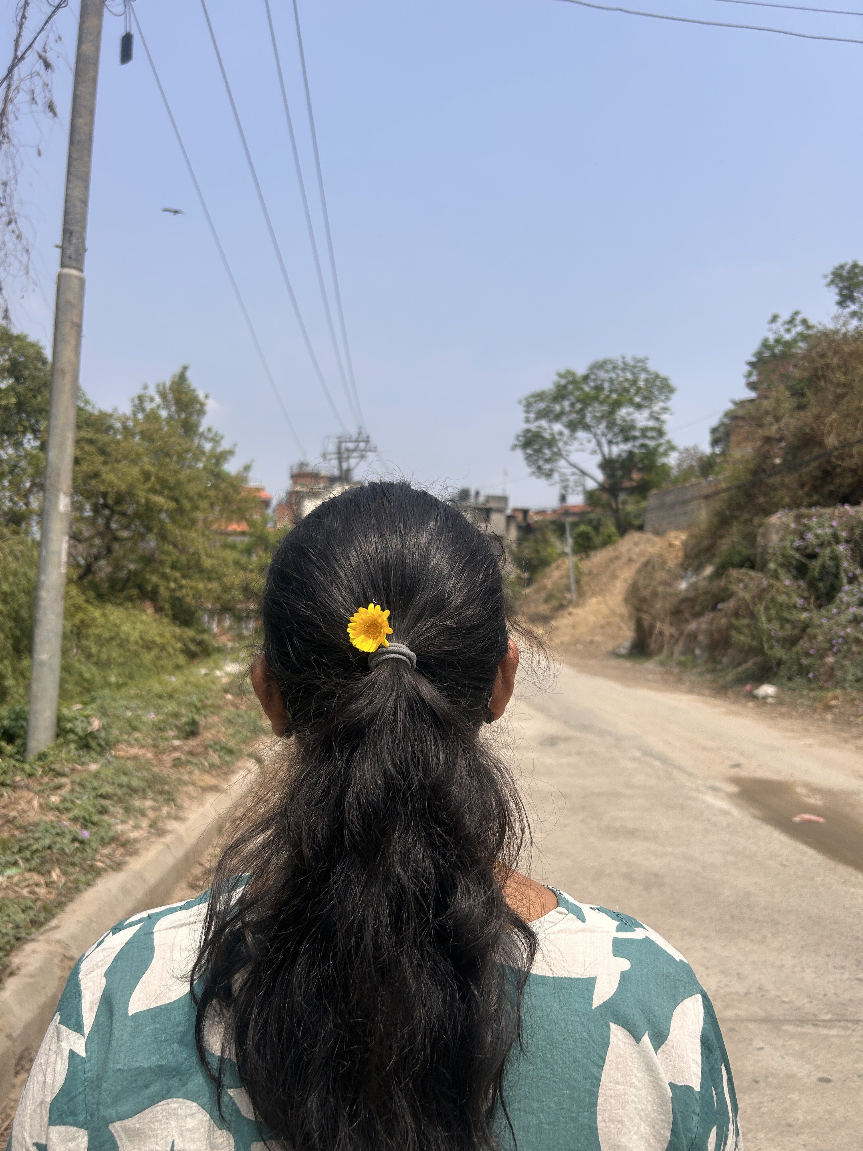 A person with long black hair tied in a ponytail, adorned with a small yellow flower, is walking down a road.