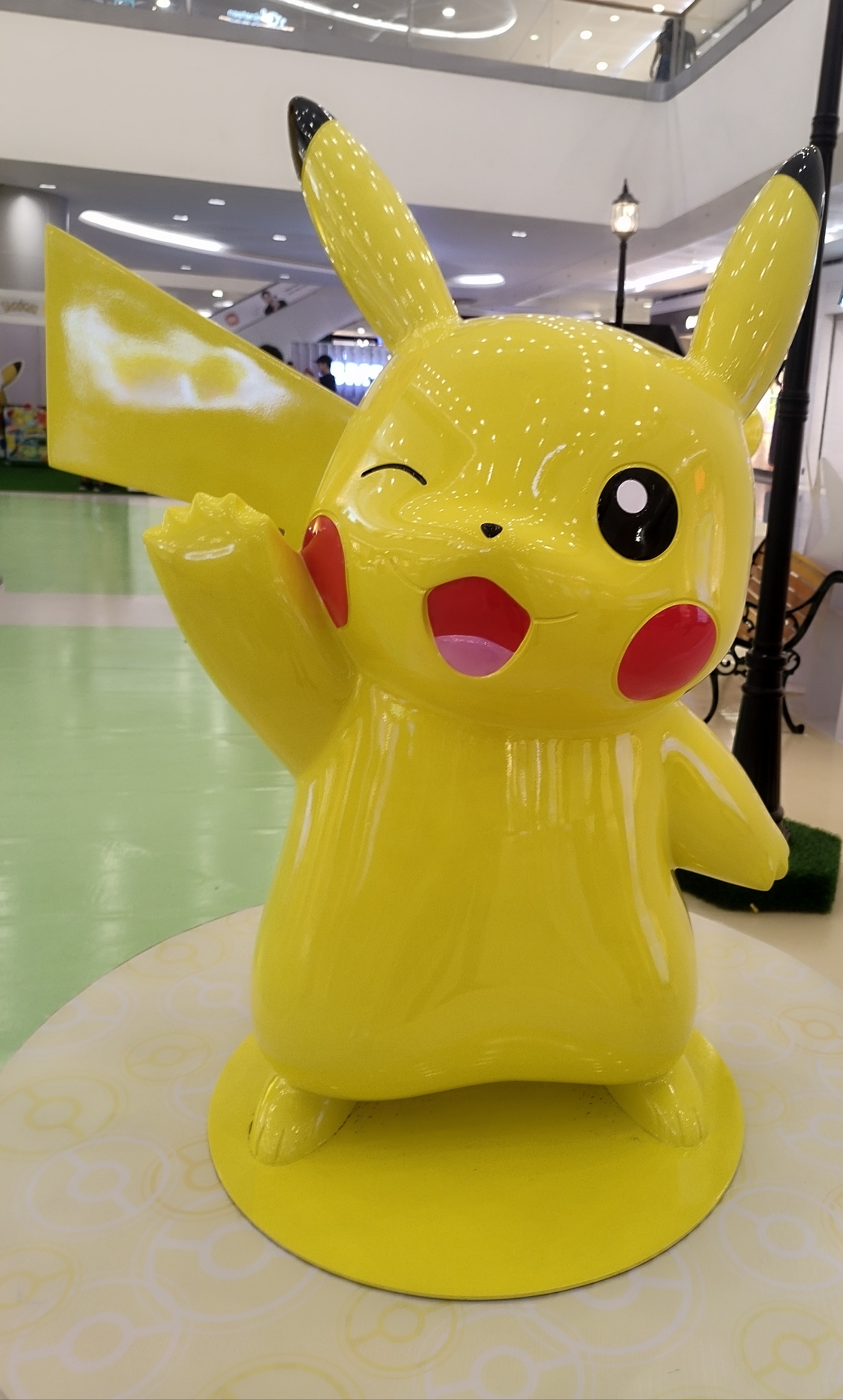 A large, cheerful Pokeeee of a yellow cartoon character with long ears, red cheeks, and a lightning bolt-shaped tail, standing on a pedestal in a shopping mall setting.