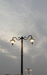 A vintage-style street lamp with two glowing lanterns against a cloudy evening sky, with more similar lamps visible in the distance.