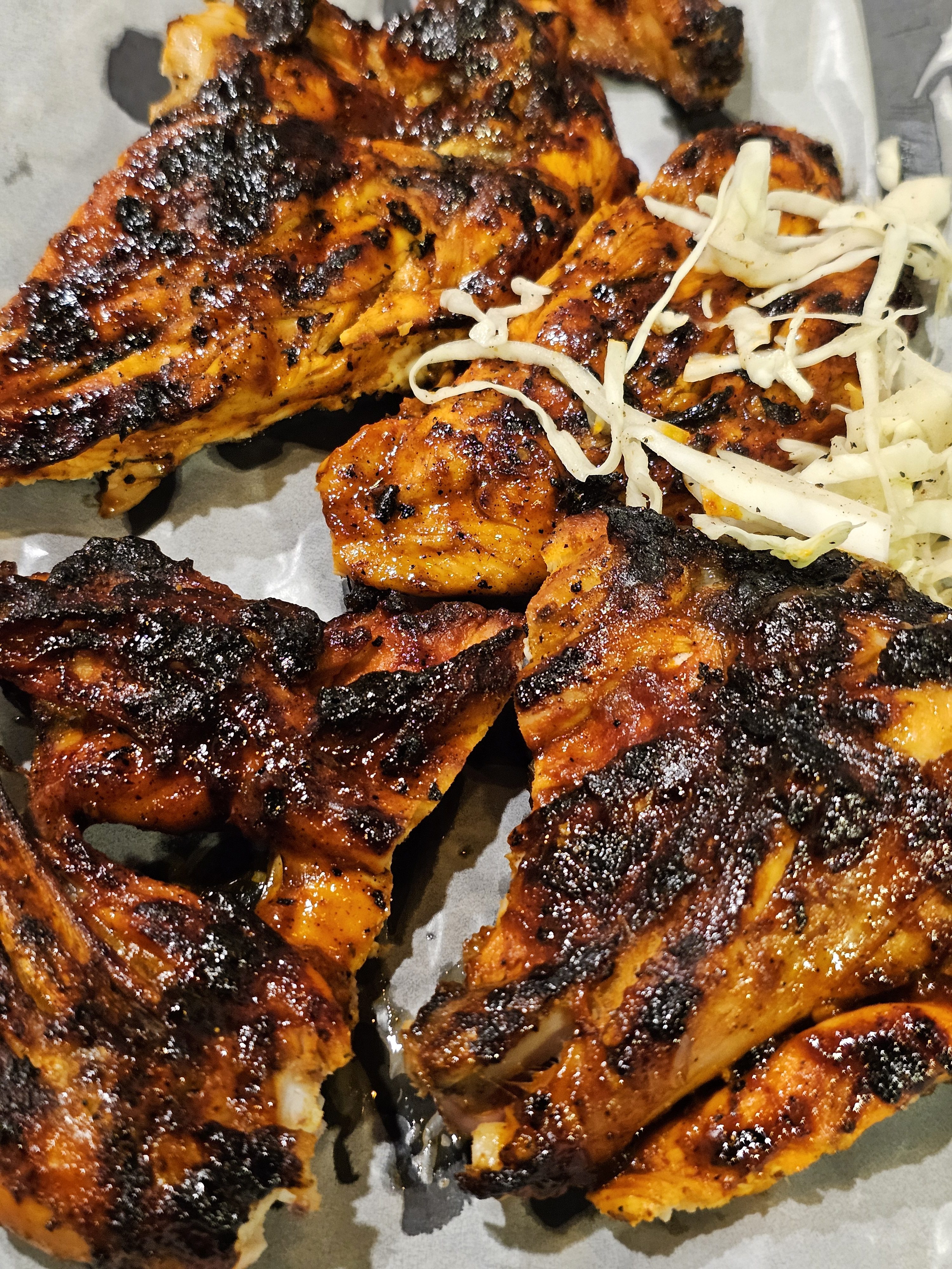A close-up of grilled chicken (Al Faham barbecued) pieces, with a slightly charred and seasoned appearance, arranged on a light-colored surface, possibly a serving plate. Shredded cabbage or lettuce is lightly scattered on the side. It was captured in Kozhikode, Kerala.
