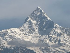 A majestic snow-covered mountain peak rises sharply against a clear blue sky, with a foreground of rugged, icy slopes. A small cloud hovers near the base of the mountain.
