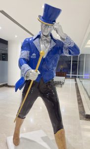 Colorful statue of a stylized man wearing a blue top hat, matching blue coat with white patterns, a silver bow tie, black pants, and yellow boots. He is holding a yellow cane and posing as if tipping his hat, placed in a well-lit indoor space with marble flooring.