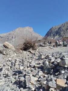 A rocky landscape with scattered stones in the foreground. In the background, there are barren mountains under a clear blue sky. A person and a horse are visible on the right side, near the base of the hillside.