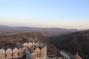 A scenic view of a fort with traditional architecture featuring arched structures, situated on a hillside. The landscape includes dry vegetation and rolling hills under a clear blue sky.