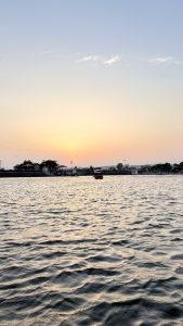 Sunset over a calm lake with gentle ripples on the water, a boat near the shore, and a lakeside area featuring small buildings, trees, and a promenade in the background.