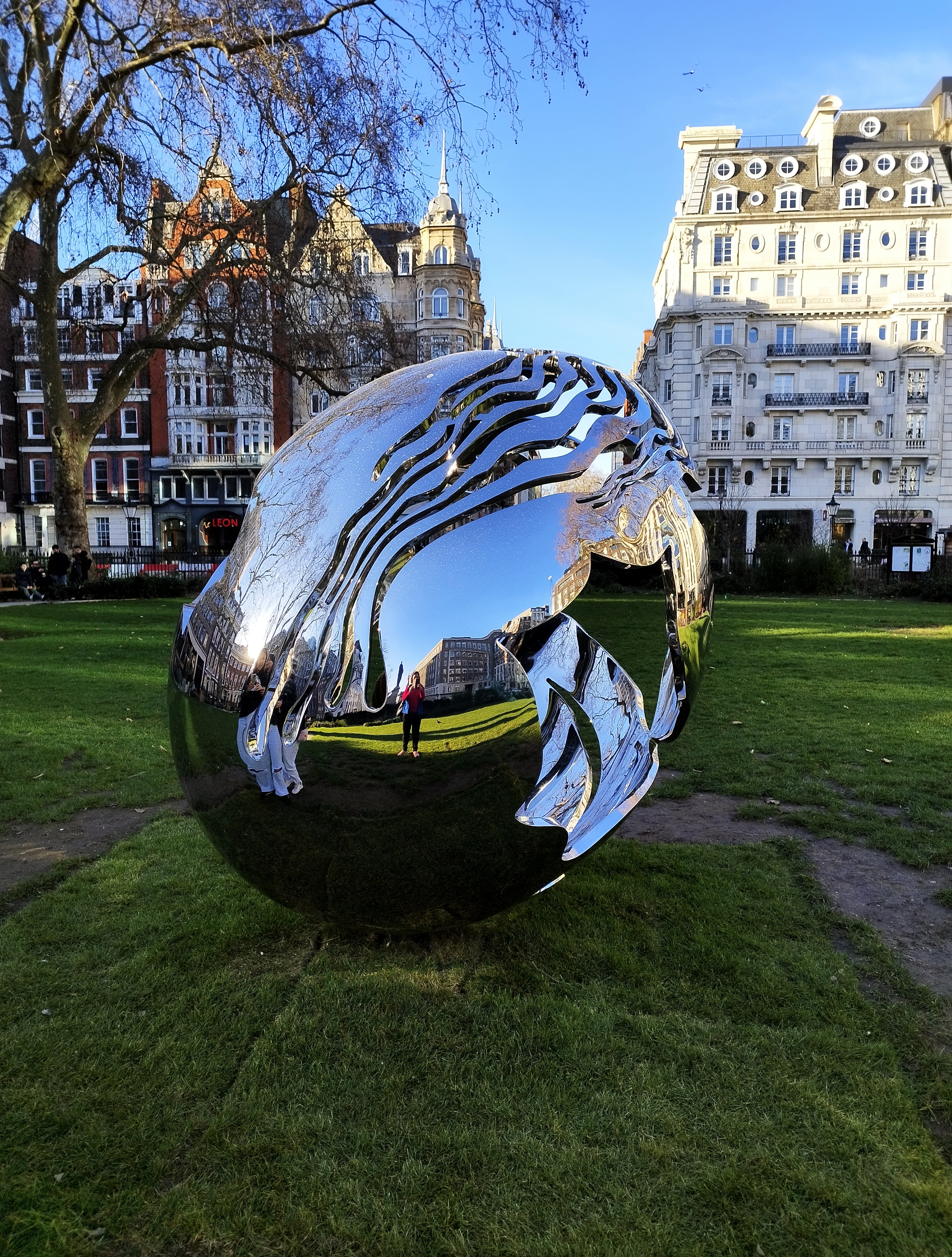 A shiny, abstract metal sculpture with reflective surfaces is situated in a grassy park area.