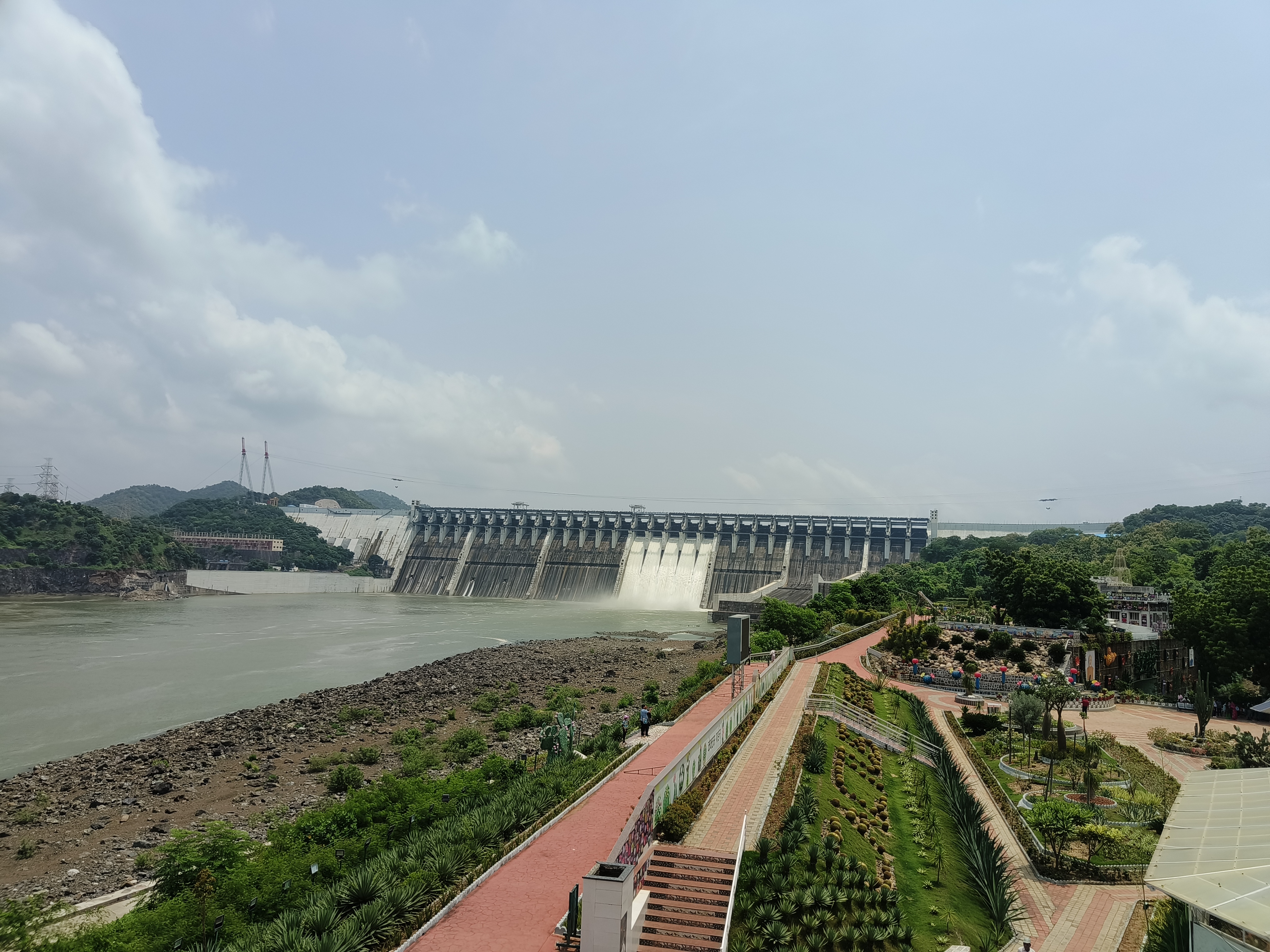 A large dam with water flowing over its spillway, surrounded by lush green vegetation. In the foreground, there is a landscaped area with pathways, plants, and decorative gardens.