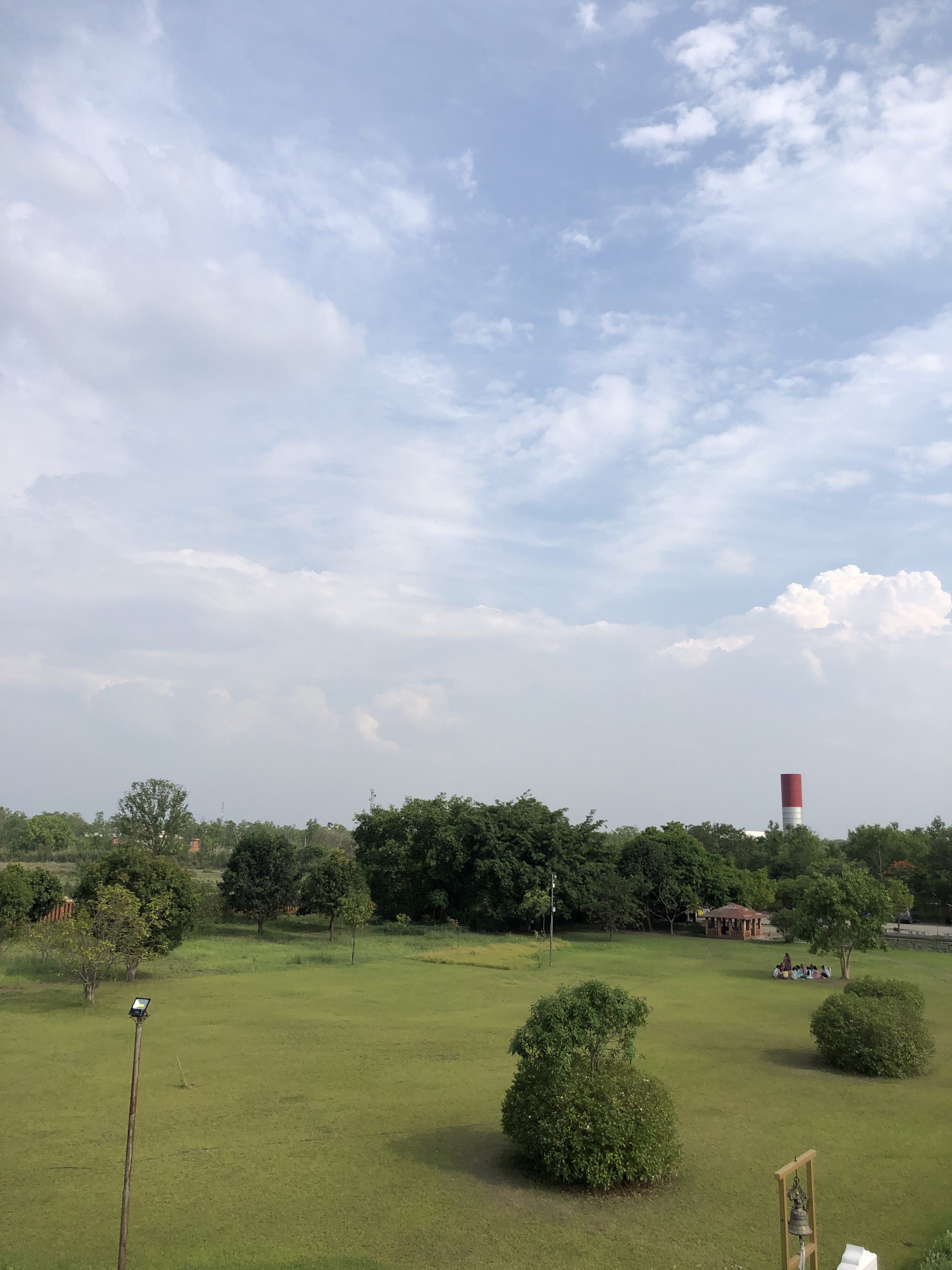 A wide grassy field with scattered bushes and trees under a cloudy blue sky. In the distance, a group of people sit near a small building and a large red and white industrial chimney is visible.