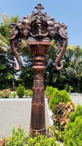 A decorative pillar with two elephant heads, set in a lush garden under a clear blue sky.
