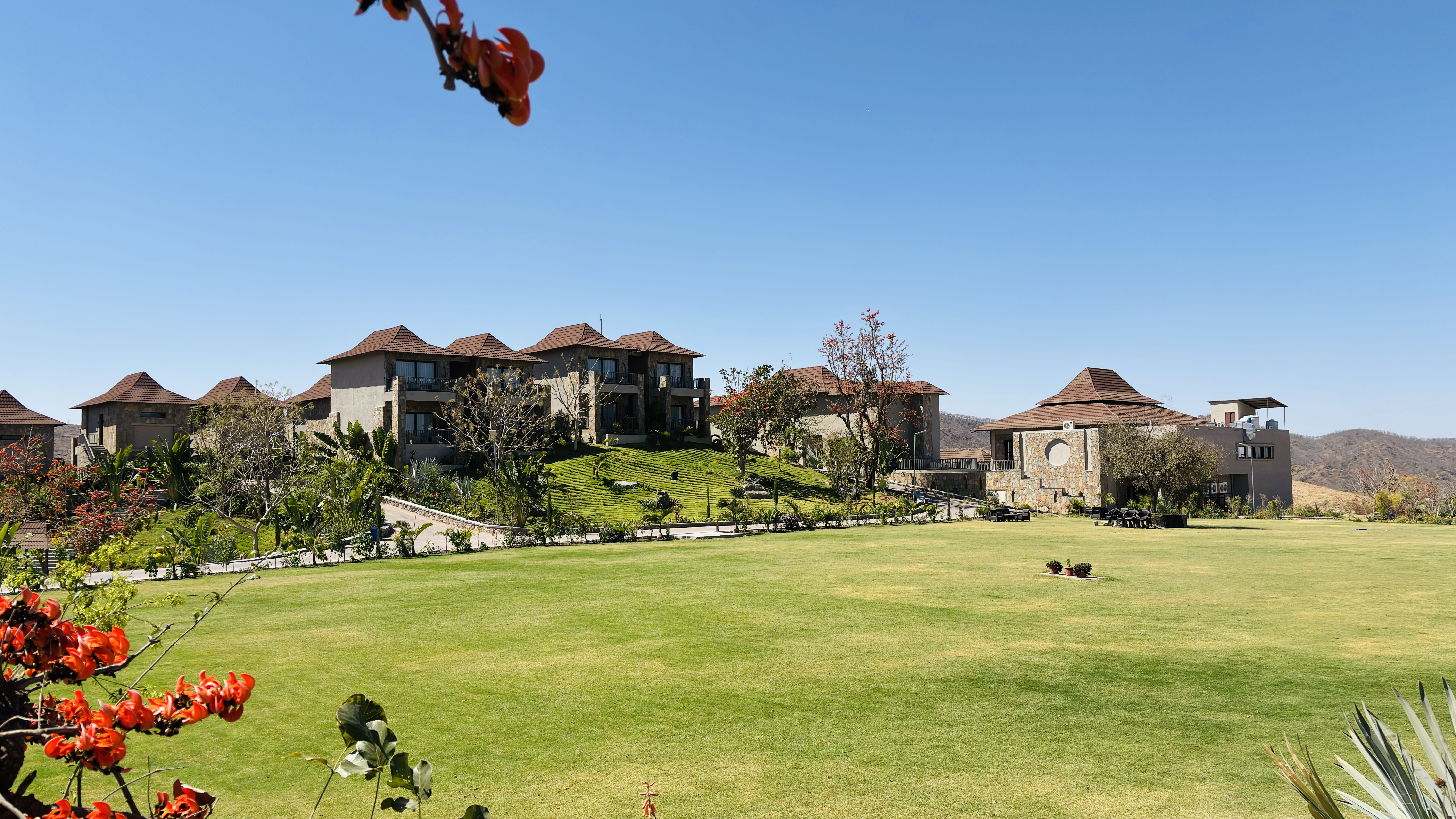 A lush green lawn with red and orange flowering plants in the foreground, leading to a group of rustic villas with sloping roofs, situated on a hillside, under a clear blue sky.