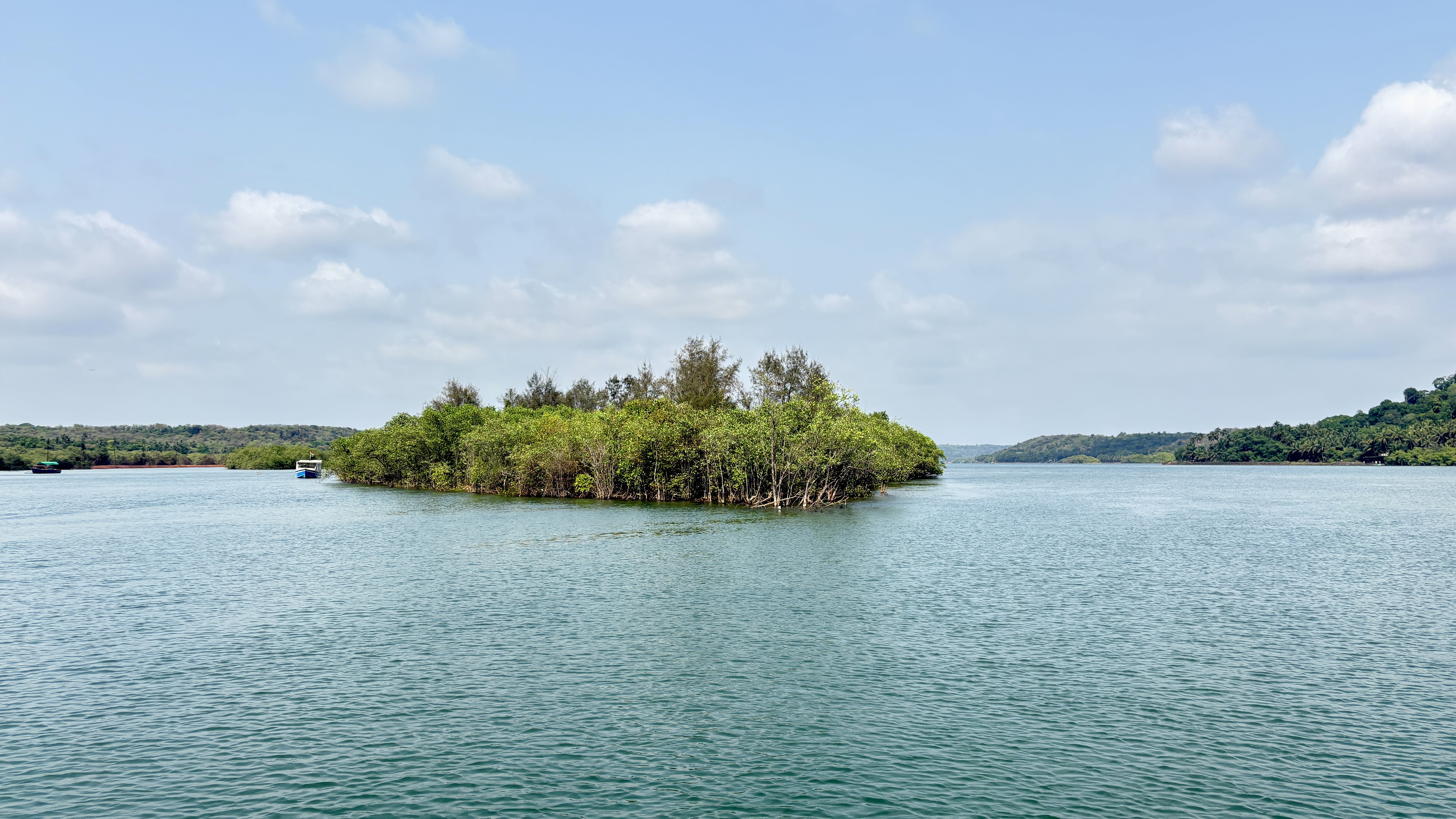 A serene view of a small, lush island surrounded by calm water, with dense green vegetation and trees. The sky is partly cloudy, and there are gentle ripples on the water's surface
