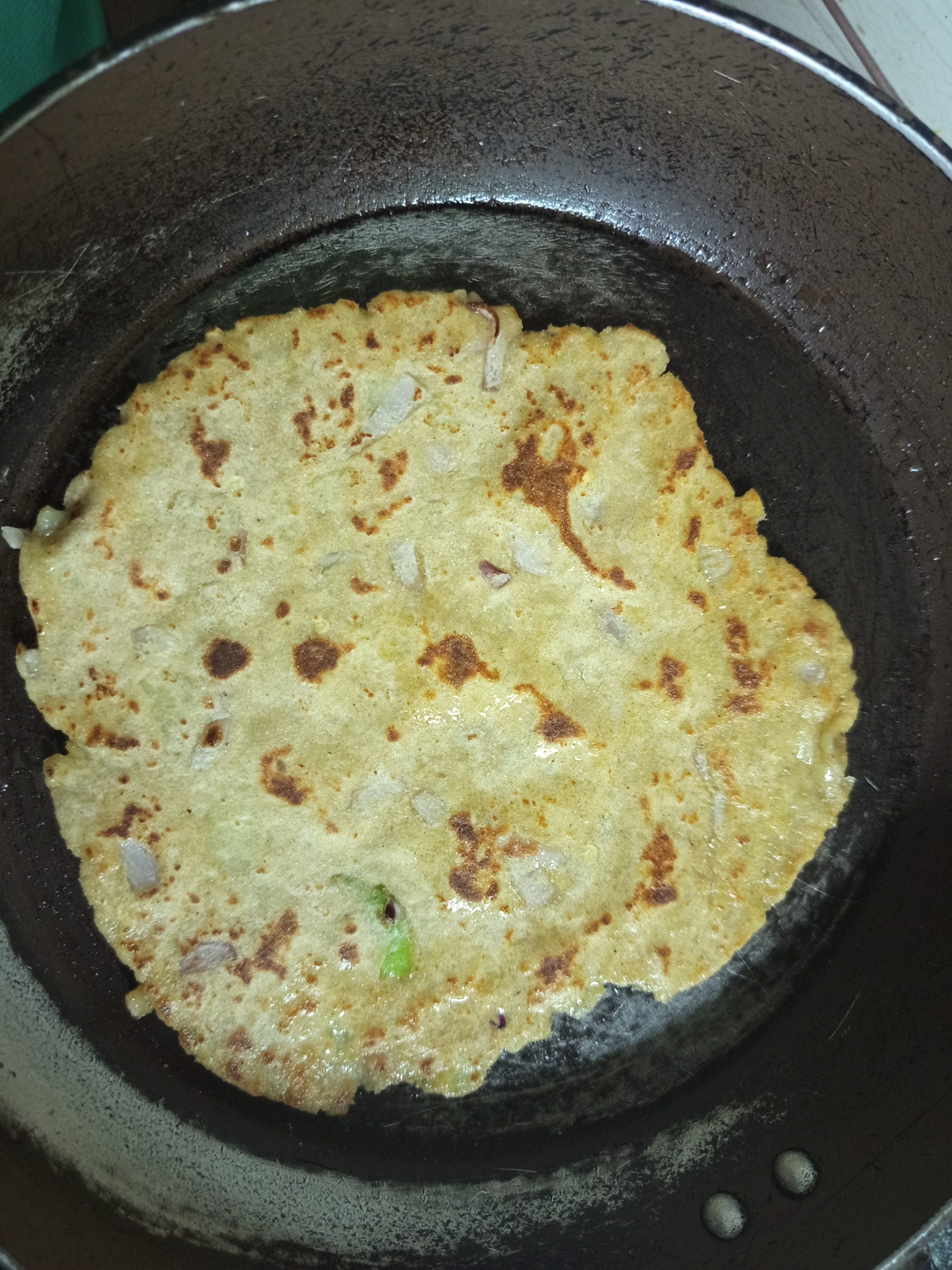 A aloo paratha with visible pieces of onions and spices is being cooked on a black pan.