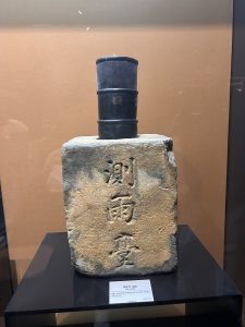 A historical rain gauge displayed in a museum. The gauge consists of a stone base with inscriptions on its front, supporting three cylindrical black containers stacked on top. The background features a neutral-toned wall, and the exhibit is placed on a glass showcase.