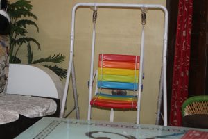 A colorful indoor swing for children with a seat featuring red, orange, yellow, green, and blue horizontal slats, suspended by a white metal frame. It's placed in a cozy room with artificial plant nearby.
