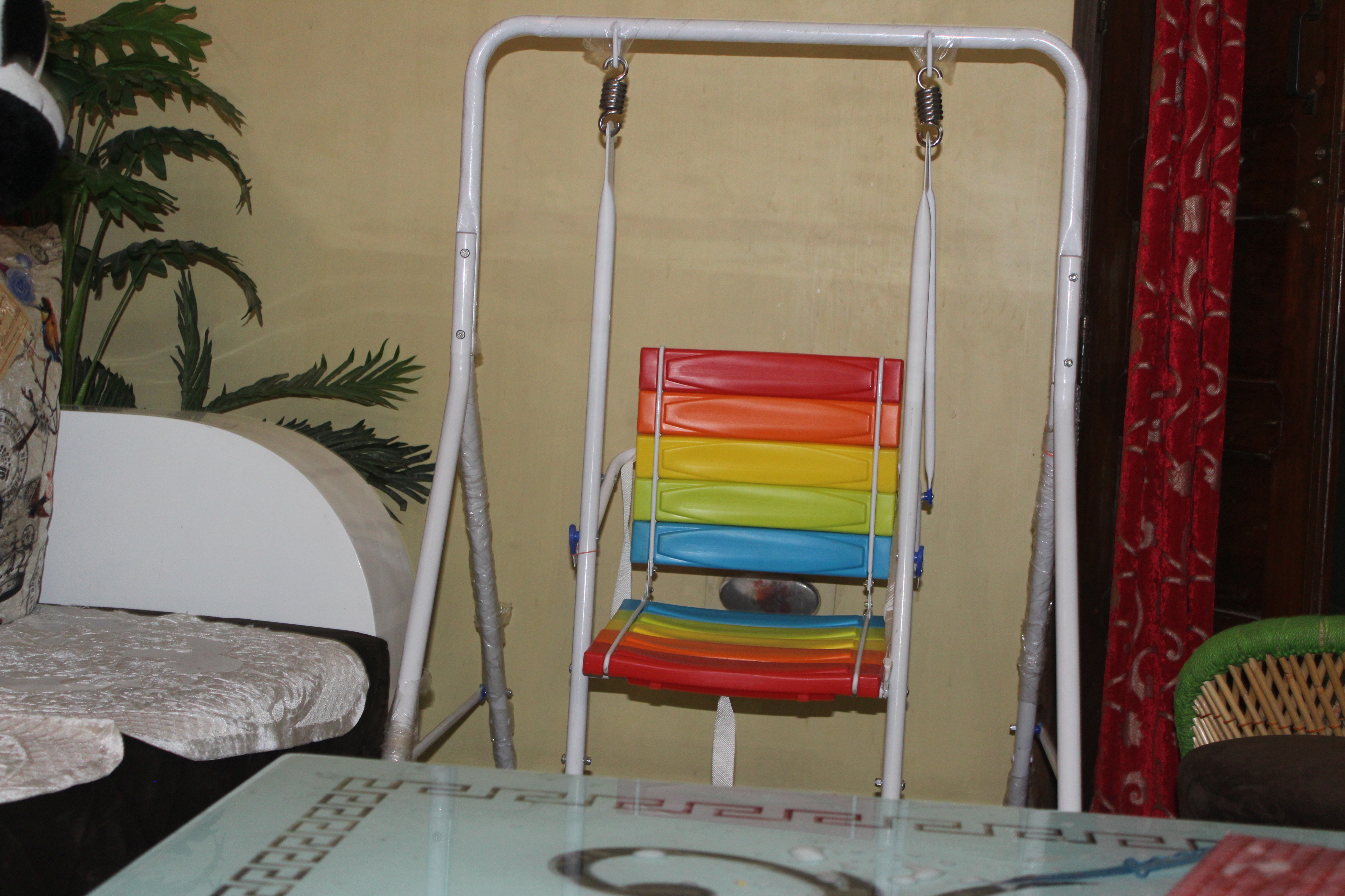 A colorful indoor swing for children with a seat featuring red, orange, yellow, green, and blue horizontal slats, suspended by a white metal frame. It's placed in a cozy room with artificial plant nearby.