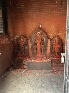 Three weathered stone statues covered in red pigment and surrounded by flowers are set against a rustic brick wall in a small, enclosed space. 