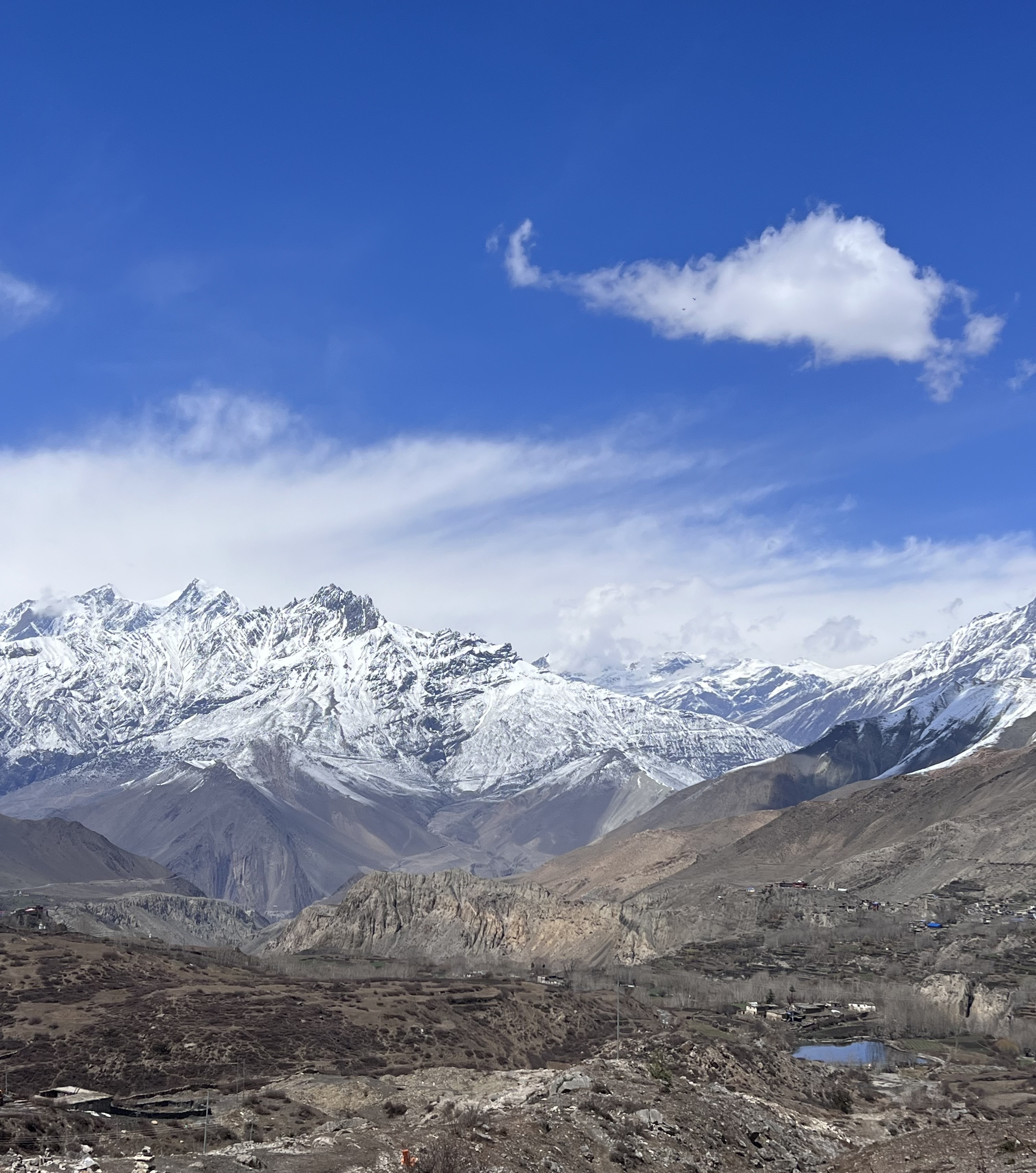 A panoramic view of snow-capped mountains under a clear blue sky with a few clouds. The foreground features rocky terrain with patches of dry vegetation and small settlements.
