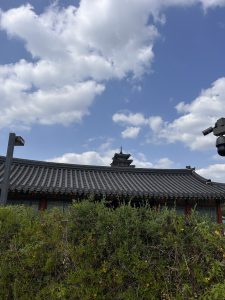 A view of a traditional Asian-style building with a curved tiled roof and ornate architecture is partially obscured by lush green bushes.