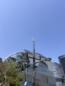 A vertical view of a street scene featuring a lamp post equipped with a small wind turbine and solar panels. In the background, modern glass buildings are visible under a clear blue sky, framed by green trees.