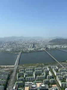 A panoramic view of a city with a river running through it, featuring high-rise buildings and residential complexes. In the foreground, several modern apartment buildings can be seen with green roofs. 