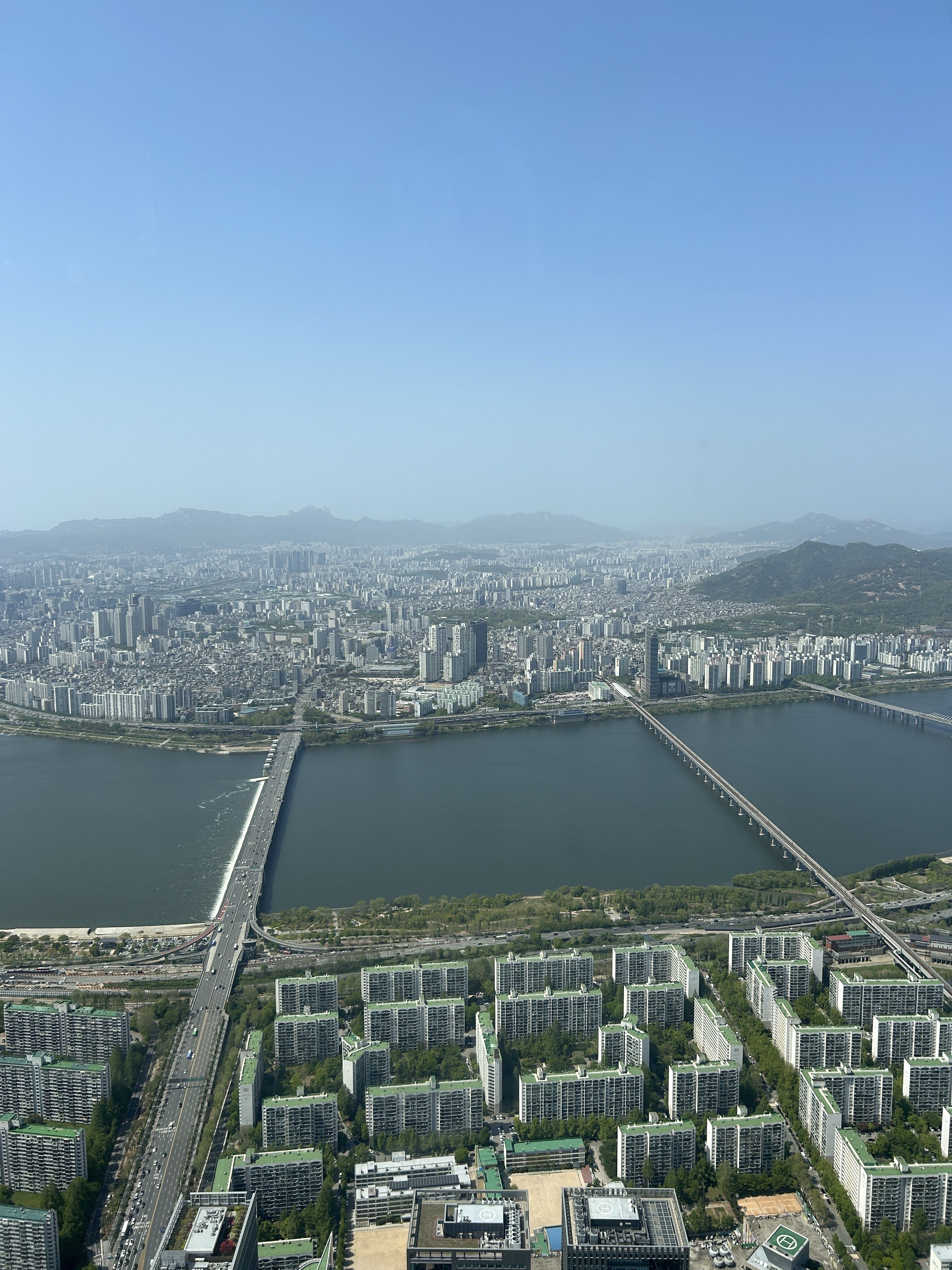 A panoramic view of a city with a river running through it, featuring high-rise buildings and residential complexes. In the foreground, several modern apartment buildings can be seen with green roofs.