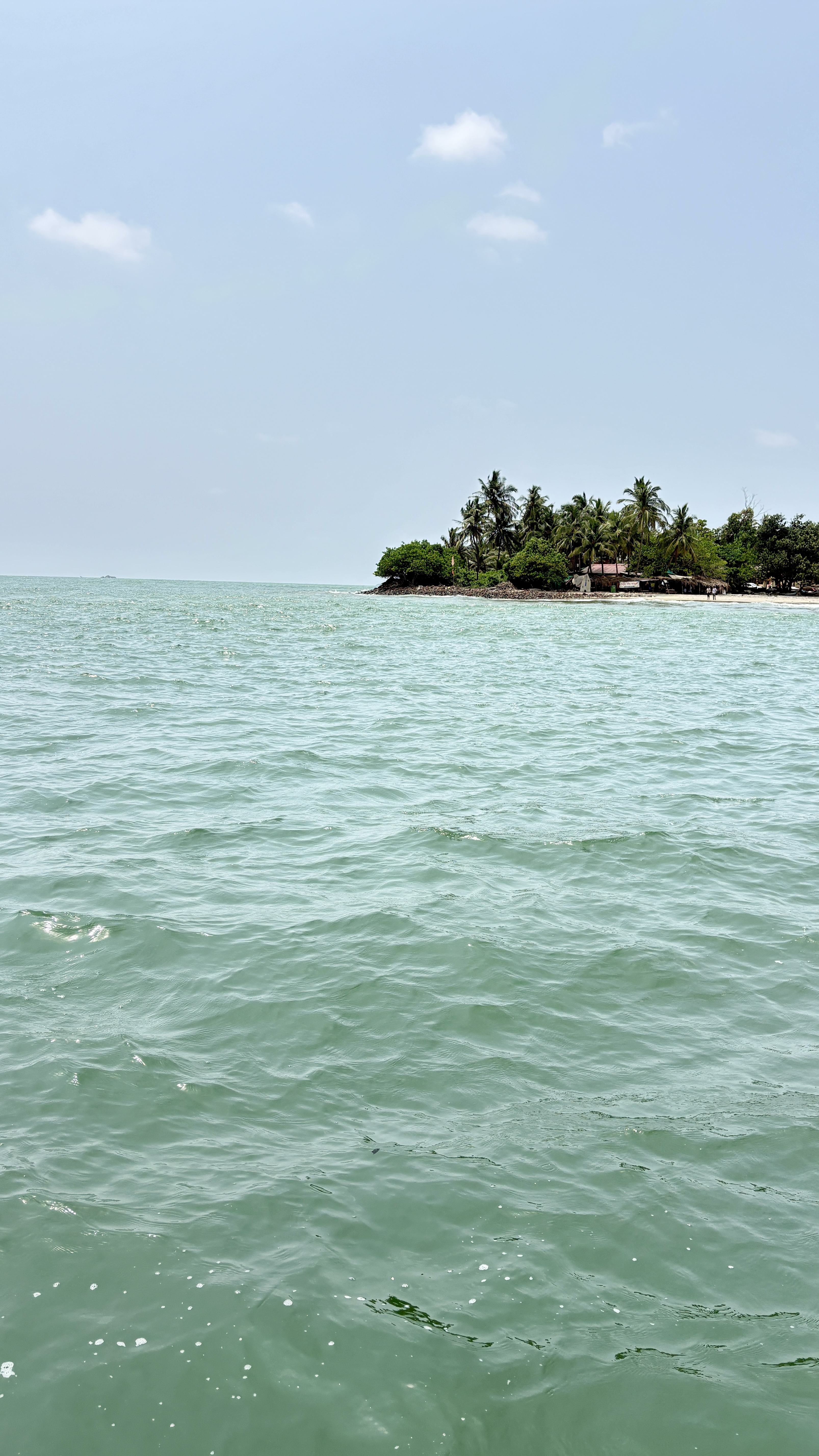 A tranquil coastal scene with light green ocean water in the foreground and a small tropical island in the distance. The island is lined with palm trees and a few small structures, under a bright, lightly clouded blue sky.