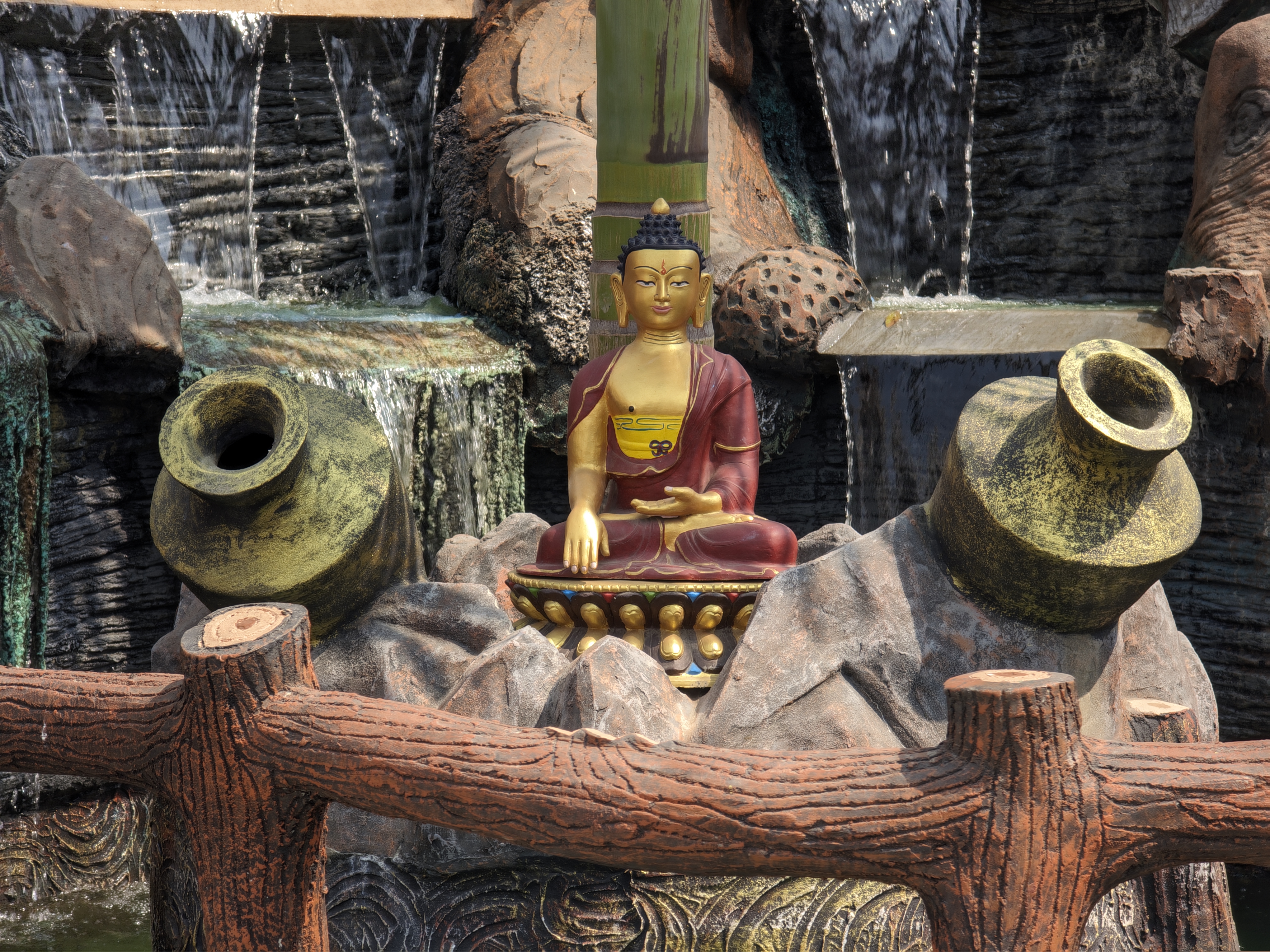 A golden Buddha statue sits in a meditation pose, centered, with two large, rough-textured pots on either side. A faux-stone waterfall backdrop enhances the serene scene, while a wooden fence-like structure stands in the foreground. #WPNP2025