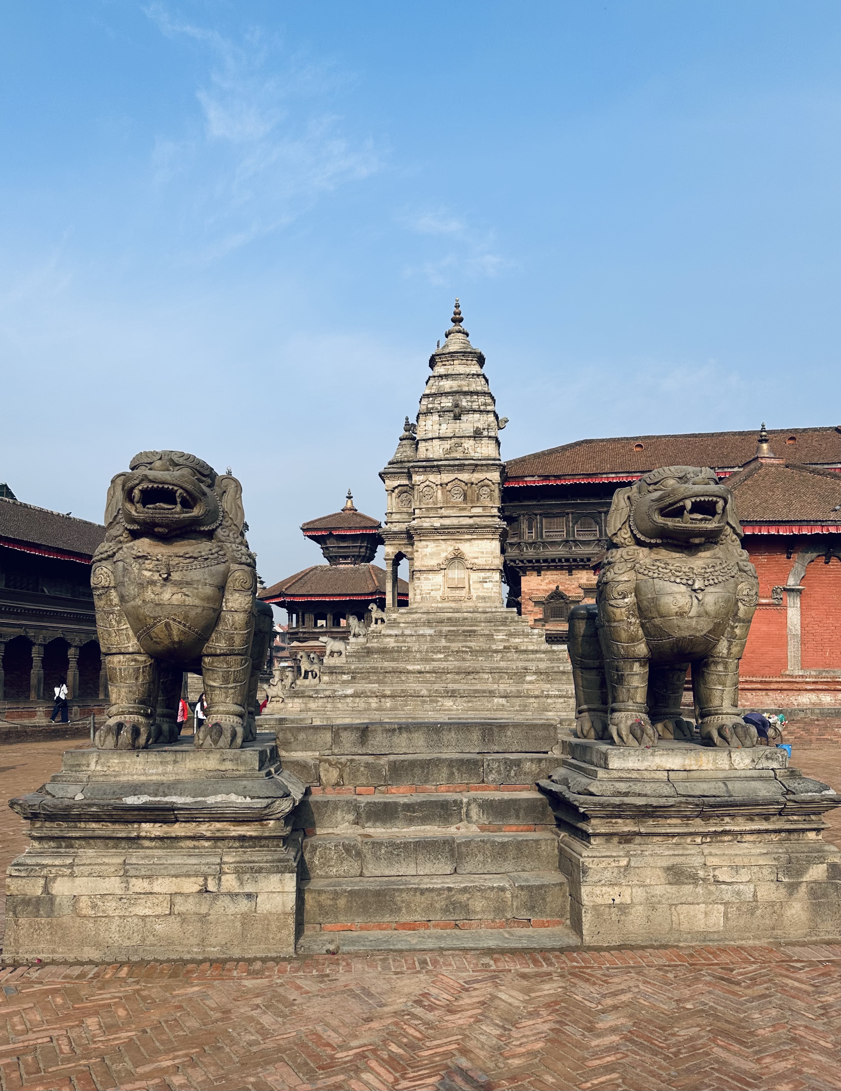 Ancient stone temple steps flanked by two large stone lion statues, leading up to a traditional temple pagoda with intricate carvings, set against a clear blue sky at Bhaktapur Durbar Square.