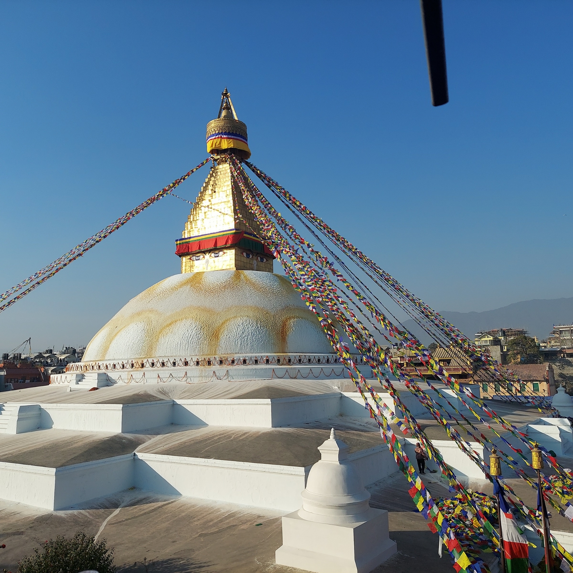 A large white stupa with a golden spire rises against a clear blue sky, adorned with colorful prayer flags extending from the top. In the background, there are distant hills and buildings