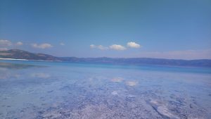 

A vast, tranquil turkey saldalake with clear blue water stretches out to meet forested mountains under a bright, cloud-dotted sky. 