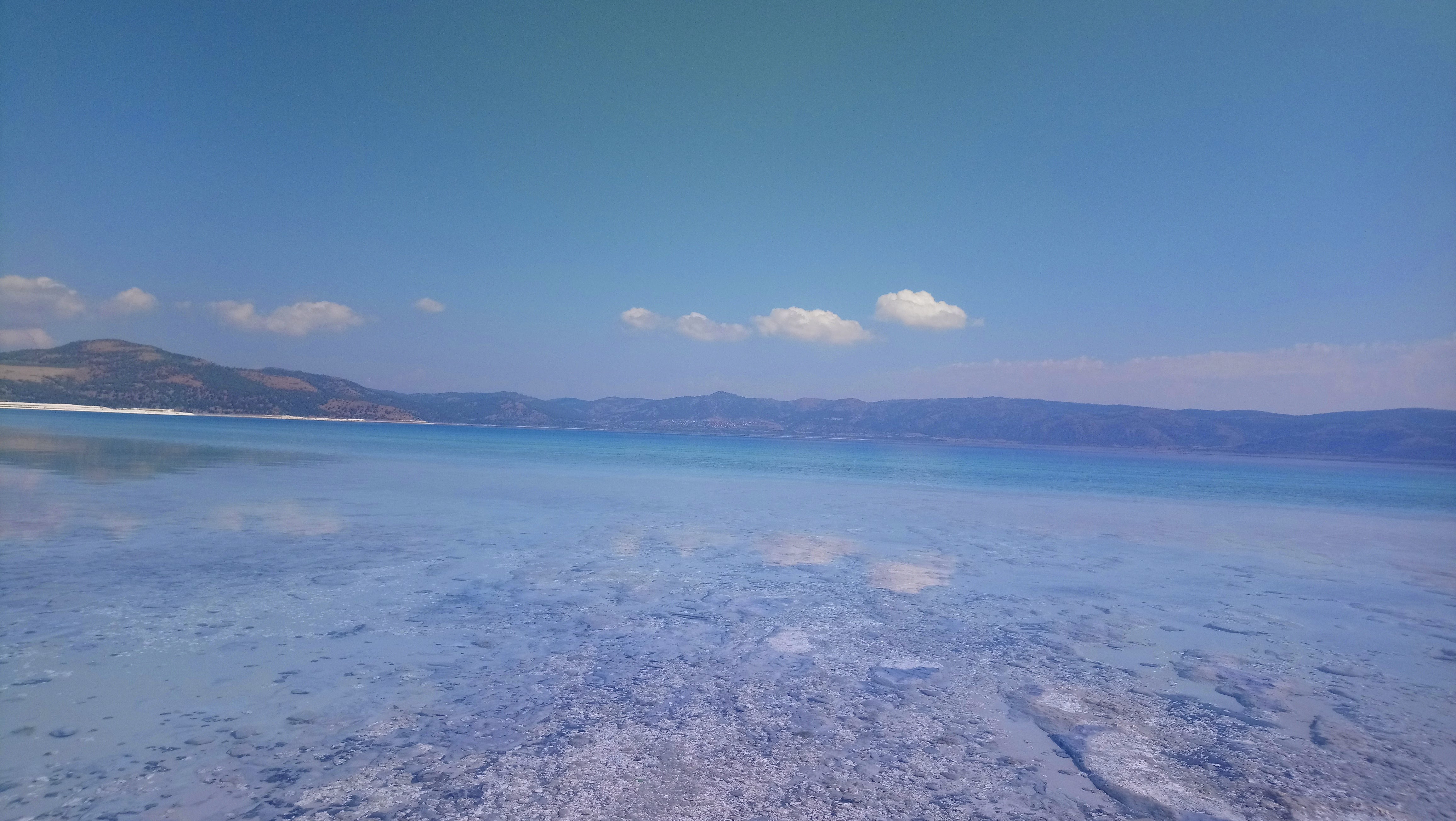 A vast, tranquil turkey saldalake with clear blue water stretches out to meet forested mountains under a bright, cloud-dotted sky.