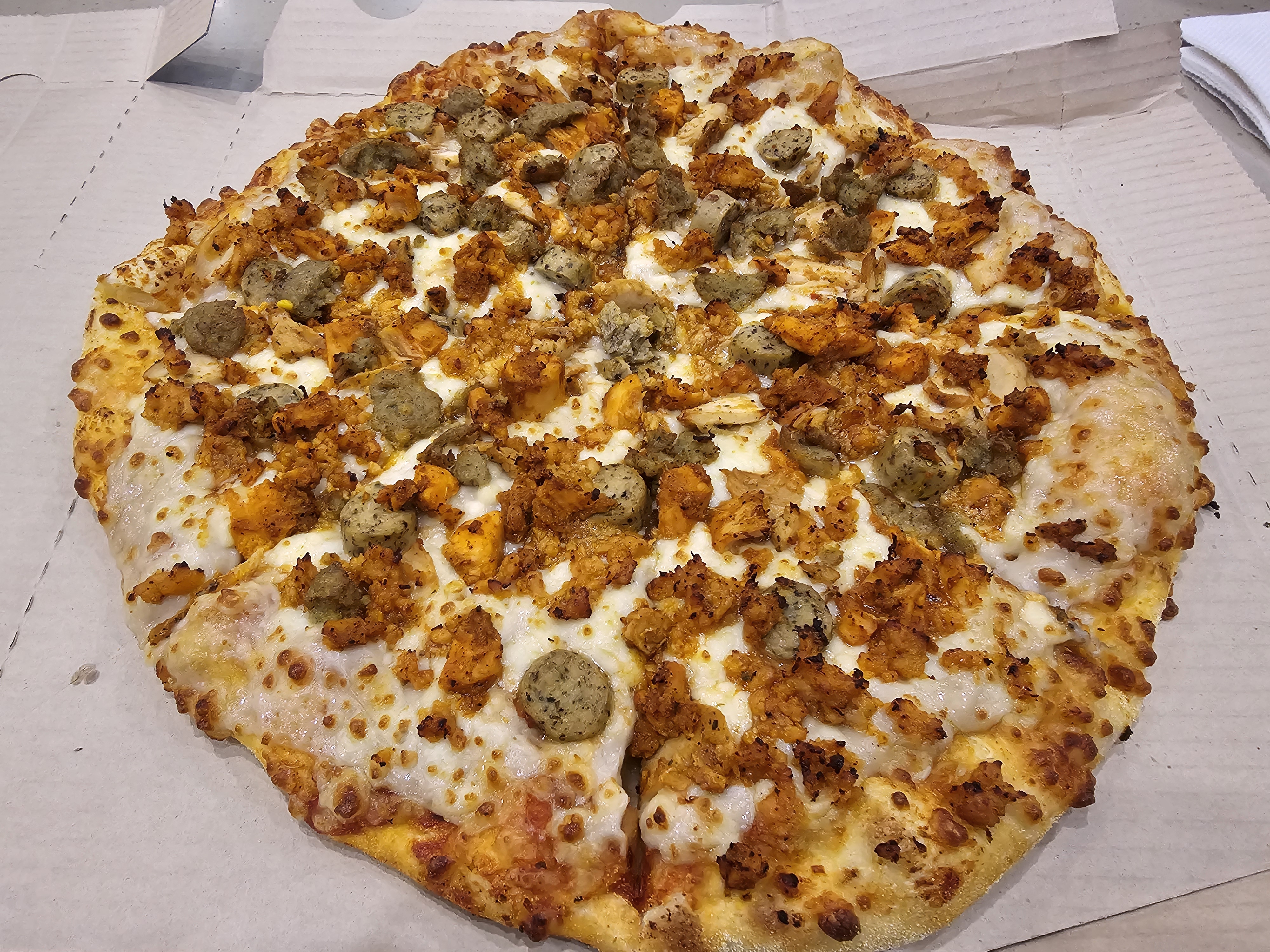 A freshly baked 5 Chicken Feast Pizza is filled with grilled Chicken, Chicken meatballs, Chicken tikka, herby chicken sausage & flavored chicken keema. The pizza is placed inside a white cardboard box. It was captured in Kozhikode, Kerala.