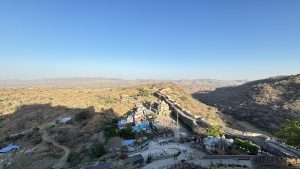 A scenic view of a sprawling ancient fort complex amidst arid, hilly terrain under a clear blue sky. The landscape includes fortified walls stretching across the hills, historical structures, and scattered greenery, with a small cluster of buildings in the foreground.
