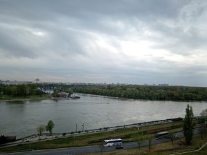 A wide view from the Belgrade Fortress shows the confluence of the Sava and Danube rivers, surrounded by greenery and urban buildings under a cloudy sky. In the foreground, there is a road with a couple of buses, while in the background, a riverboat is docked near a small pier. Trees line the riverbanks, and the landscape features a mix of residential buildings in the distance.