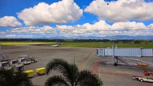 A view of an airport runway under a blue sky filled with fluffy clouds. In the foreground, there is a boarding gate with a staircase leading down, and several ground service vehicles including luggage carts and tugs.