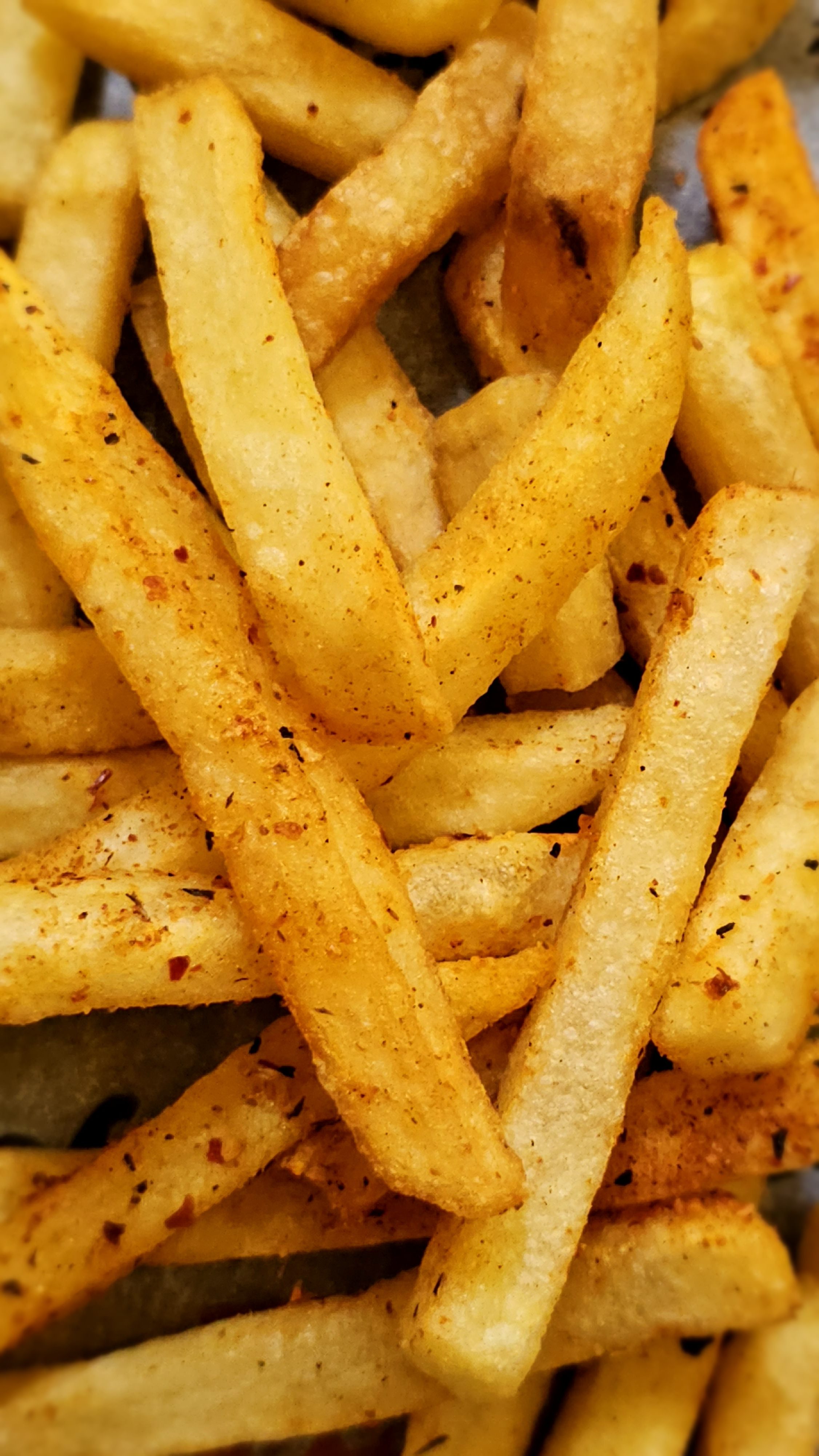 A close-up image of golden-brown French fries, seasoned with spices, arranged in a haphazard pile. Some fries are thicker, while others are thinner, and they exhibit a crispy texture with visible seasonings on their surface.