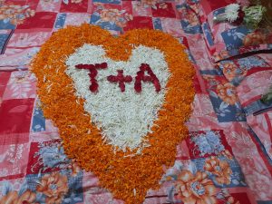 A heart shape made of orange and white flower petals is arranged on a floral patterned bedspread. The initials “T + A” are formed in the center with red petals.
